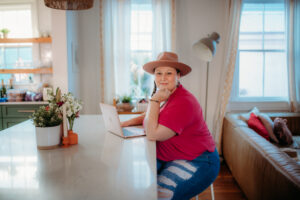 Meagan Hernandez is smiling at the camera while sitting at the counter on her computer. She's wearing a red top and ripped jeans, sitting on a stool. Her hair is in a single braid on the right side of her face, and she's wearing a brown wide brimmed hat.