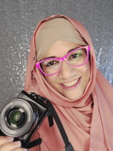 Shehla Fraizi is smiling at the camera and holding a camera. She's wearing hot pink glasses and a tan and light pink hijab. The background is a sparkly silver sequins.