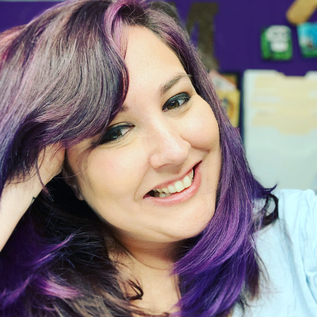 Molly Ryden is smiling at the camera and leaning her head onto her hand. Black and purple hair falls across her hand and shoulders. Her top is blue.
