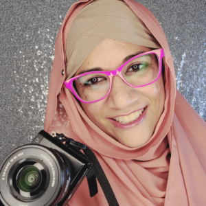 Shehla Fraizi is smiling at the camera and holding a camera. She's wearing hot pink glasses and a tan and light pink hijab. The background is a sparkly silver sequins.