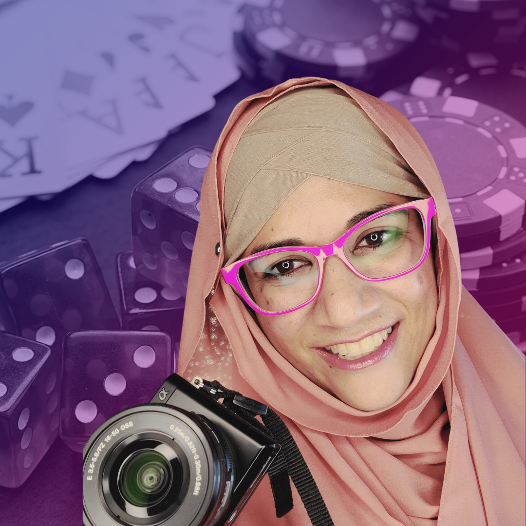 Shehla Fraizi is smiling at the camera and holding a camera. She's wearing hot pink glasses and a tan and light pink hijab. The background is a casino table with cards, dice, and chips.