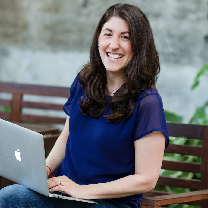 Bev Feldman is sitting on a brown bench outside and smiling at the camera as she works on her laptop. She's wearing jeans and a royal blue flowy top.