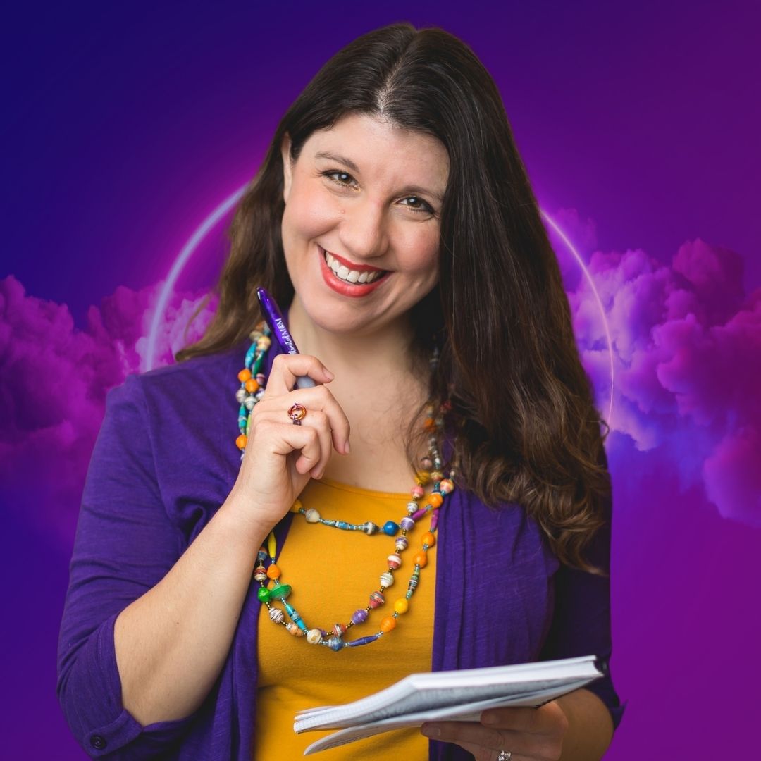 Valerie Friedlander is smiling while holding a pen raised to her chin and a notebook. She is wearing an orange top with a purple cardigan and a necklace of colorful beads.