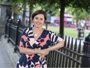 Louise Brogan stands outside, leaning against an wrought iron fence. She's wearing a Blue, Coral, and Pink top and a beautiful silver statement necklace.