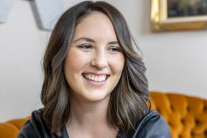 Melanie Scroggins is smiling and looking past the camera. She's wearing a dark hoodie and sitting on an orange couch.