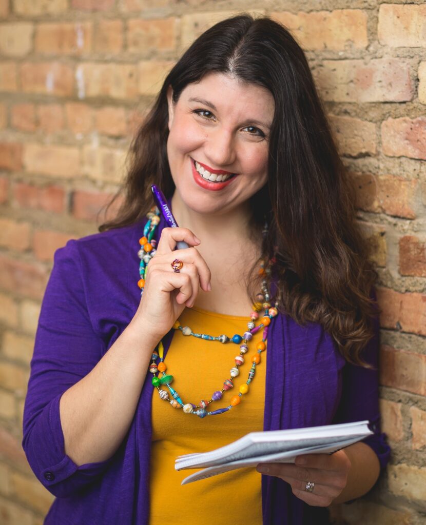 Valerie Friedlander is smiling while holding a pen raised to her chin and a notebook. She is wearing an orange top with a purple cardigan and a necklace of colorful beads.