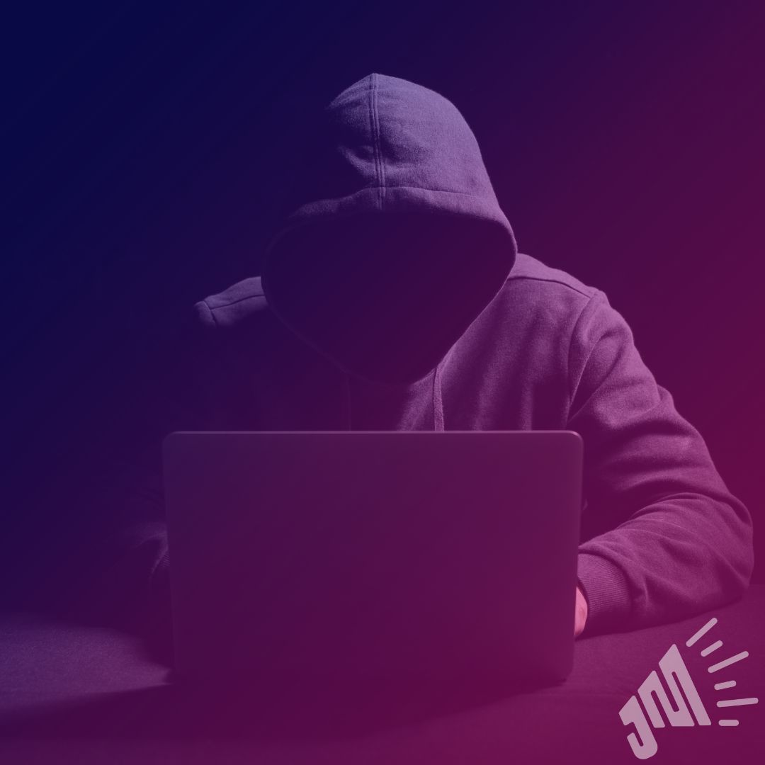 A person wearing a hoodie is in front of a laptop. The persons face is completely hidden in the shadows and the hood.