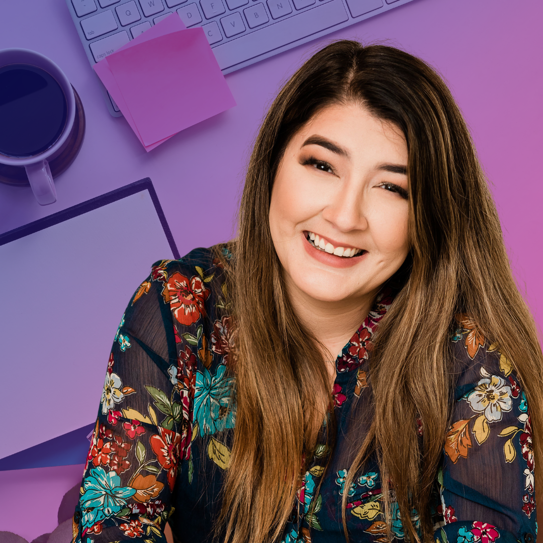 Emilie Given is smiling and her long brown hair flows over a blue sheer top with embroidered flowers in blue, orange, red, white, and green. The background is an office flatlay with a keyboard, post-it notes, coffee cup, and tablet.