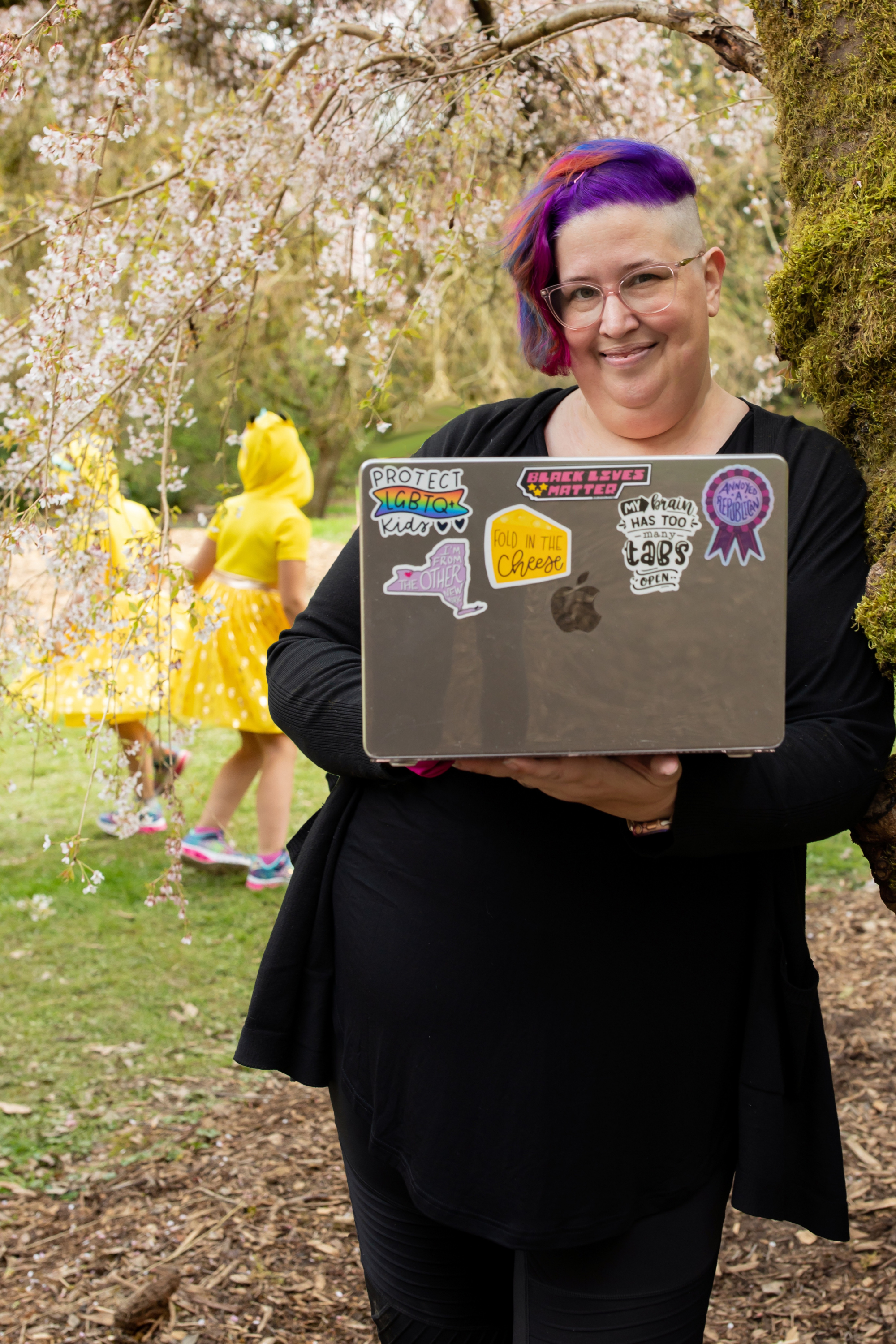 Meg is smiling, leaning against a mossy tree and holding her laptop. Behind her, her youngest 2 kids, in Pikachu dresses, dance in the cherry blossoms.
