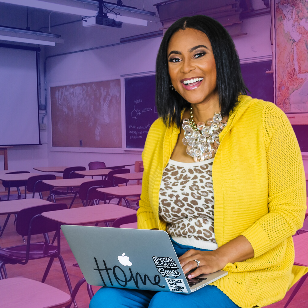 Cindy Lumpkin is smiling and wearing a yellow cardigan over a leopard print top with a gorgeous necklace. She has her laptop on her lap. The background is an empty school classroom.
