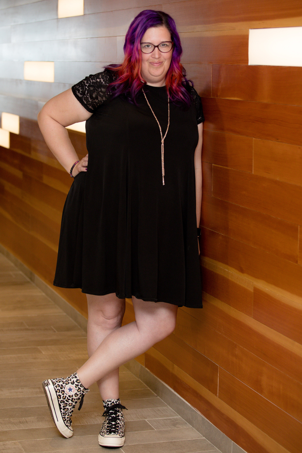 Meg is smiling and leaning against a wall. She's wearing a black dress with lace sleeves, a long gold necklace, and showing off leopard print converse high tops. Her hair reminds me of the Instagram logo in purple, pink, and orange.