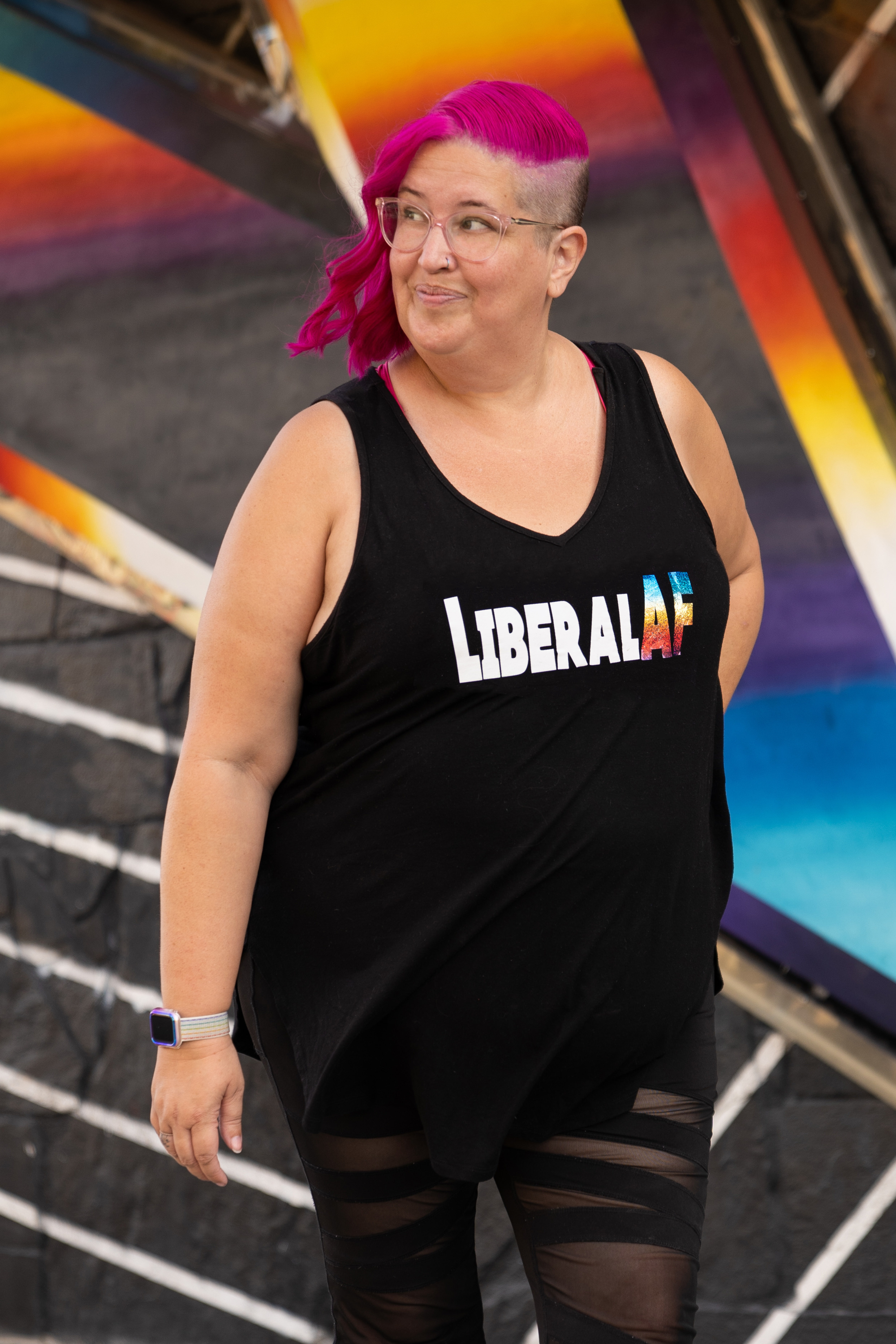 Meg is smirking as she stands in front of an edgy urban wall. She's looking up and to the left. Her hair is pink and undercut. "Liberal AF" is written across her shirt.