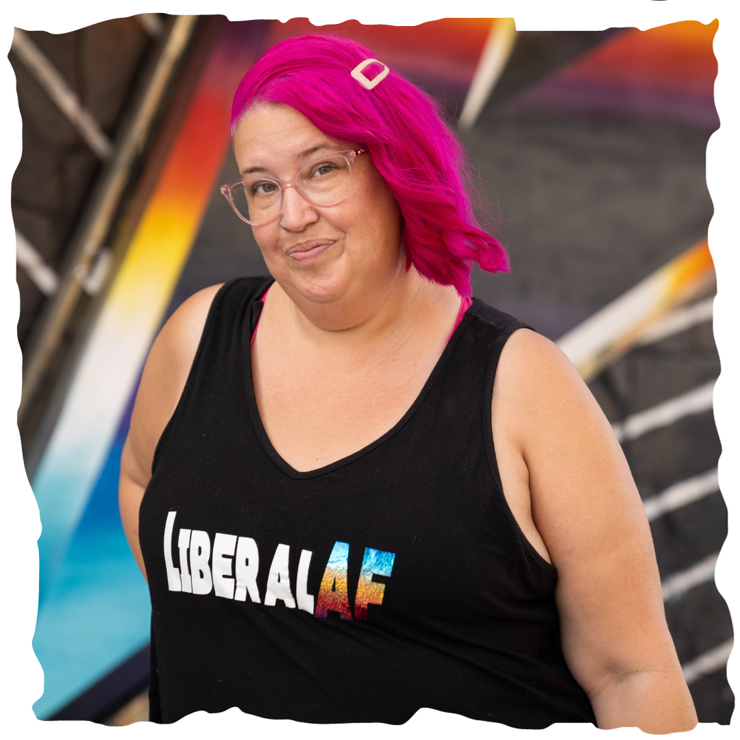 Meg is smiling at the camera. Her hot pink hair is captivating. Her black top says "Liberal AF" and the background is edgy and urban.