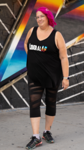 Meg is standing confidently in front of an edgy urban building. Her hot pink hair is pulled to one side, and her top reads "Liberal AF" she's also wearing athletic capris with some sexy mesh cutouts and some comfy sneakers.