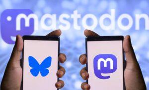 Two hands each hold a cell phone. On the left the phone displays the Bluesky logo. On the right, the phone displays the Mastodon logo. The background is blurry with the mastodon logo.