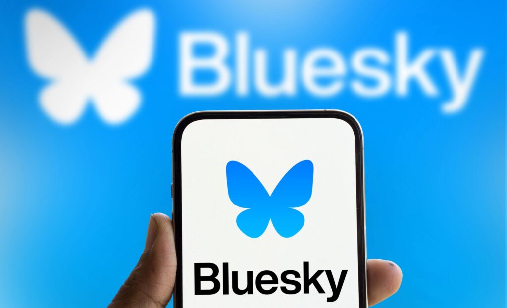 A hand holds a cell phone displaying the Bluesky icon and logo. The background is light blue and also displays the full logo of Bluesky, slighty blurred.