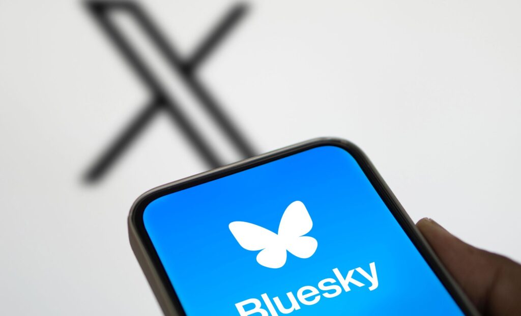 The Bluesky app logo is pulled up on a cell phone. In the background the X logo is blurred out.