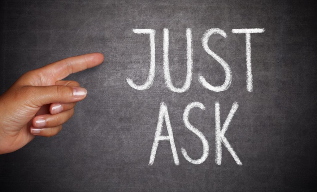 "Just Ask" is written in chalk on a chalkboard. A finger is pointing at the words.