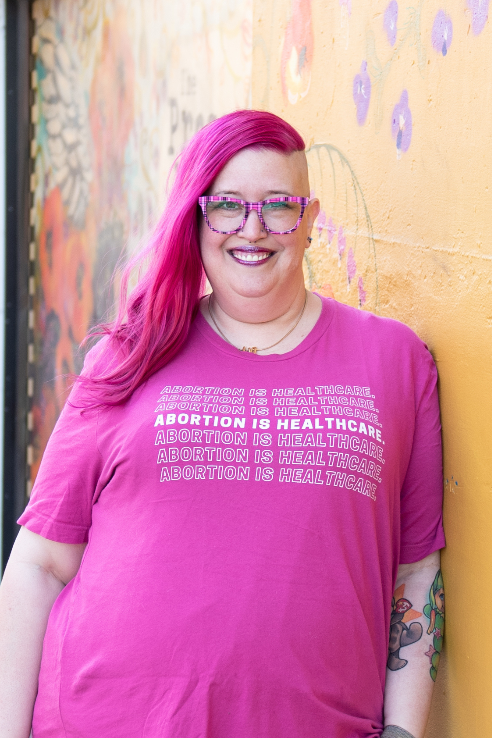 Meg Brunson, with long bright pink hair and shaved sides smiles while standing in front of glass blocks. They wear pink plaid glasses and a matching pink shirt that repeatedly states in white text, "Abortion is Healthcare." They have pink glitter lipstick and a nose piercing.