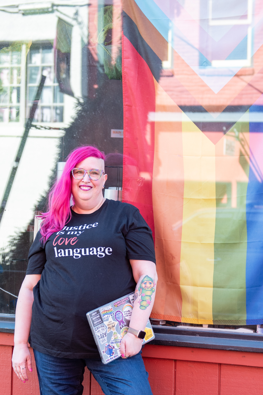 Meg Brunson, with long bright pink hair and shaved sides stands in front of a window displaying a Progress Pride flag. They wear colorful glasses, a black shirt reading “Justice is my love language,” and hold a laptop covered in colorful stickers. They smile confidently, with a tattoo of a cartoon-style glow worm visible on their forearm.