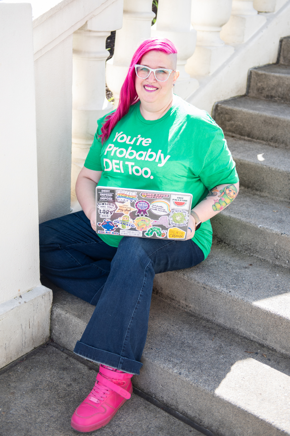 Meg Brunson, with long bright pink hair and shaved sides sits on outdoor steps, smiling and holding a sticker-covered laptop. They wear white-framed glasses, a green shirt that says "You're Probably DEI Too.", dark jeans, and bright pink shoes. A colorful glow worm tattoo is visible on their forearm.